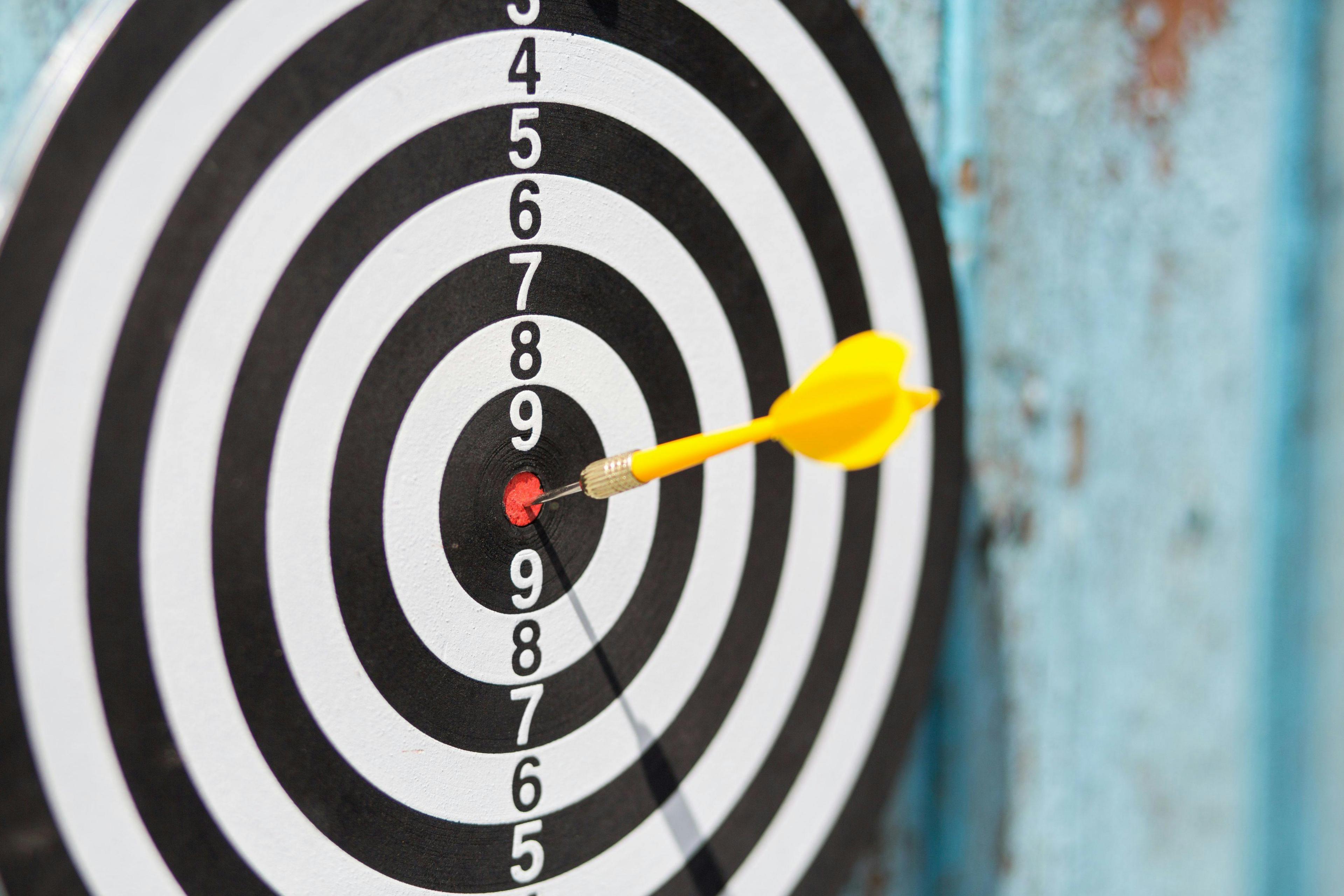 a yellow dart that has hit the bullseye on a black and white circular dartboard. The dart is lodged in the red center, and the rings on the board are numbered, with the outermost circle displaying the number 10, and the inner circles marked down to the number 9 around the bullseye. The dartboard is mounted on a vertical surface.