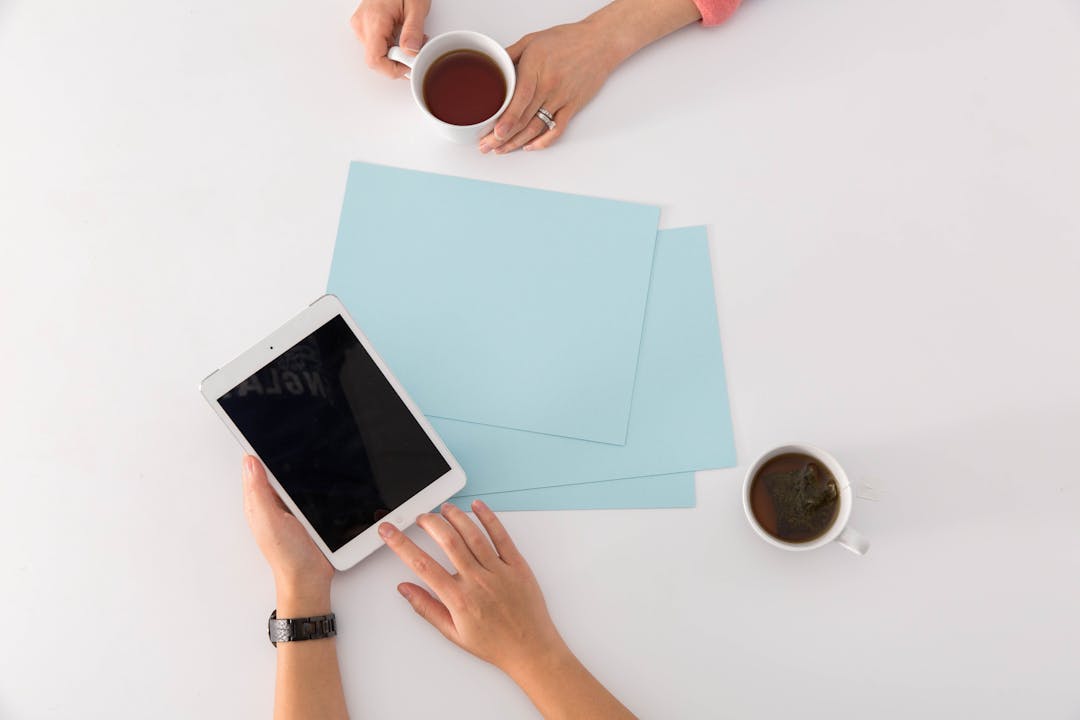 blue sheets of paper on a surface with 2 cups of tea and hands holding a tablet