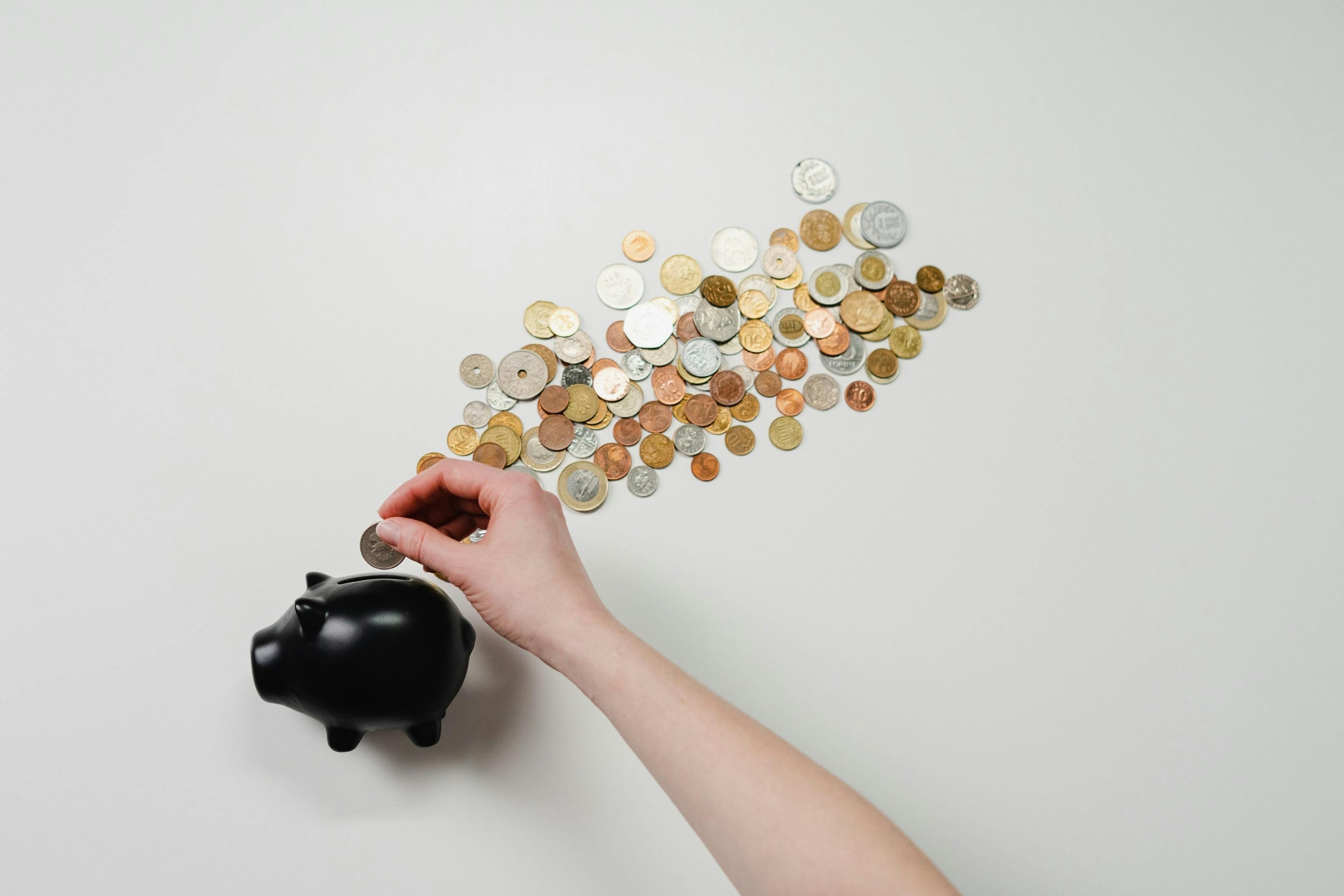 A small, black piggybank on a white background, with coins of various shapes and sizes floating in a cloud above it. A hand is putting another coin in the piggybank.