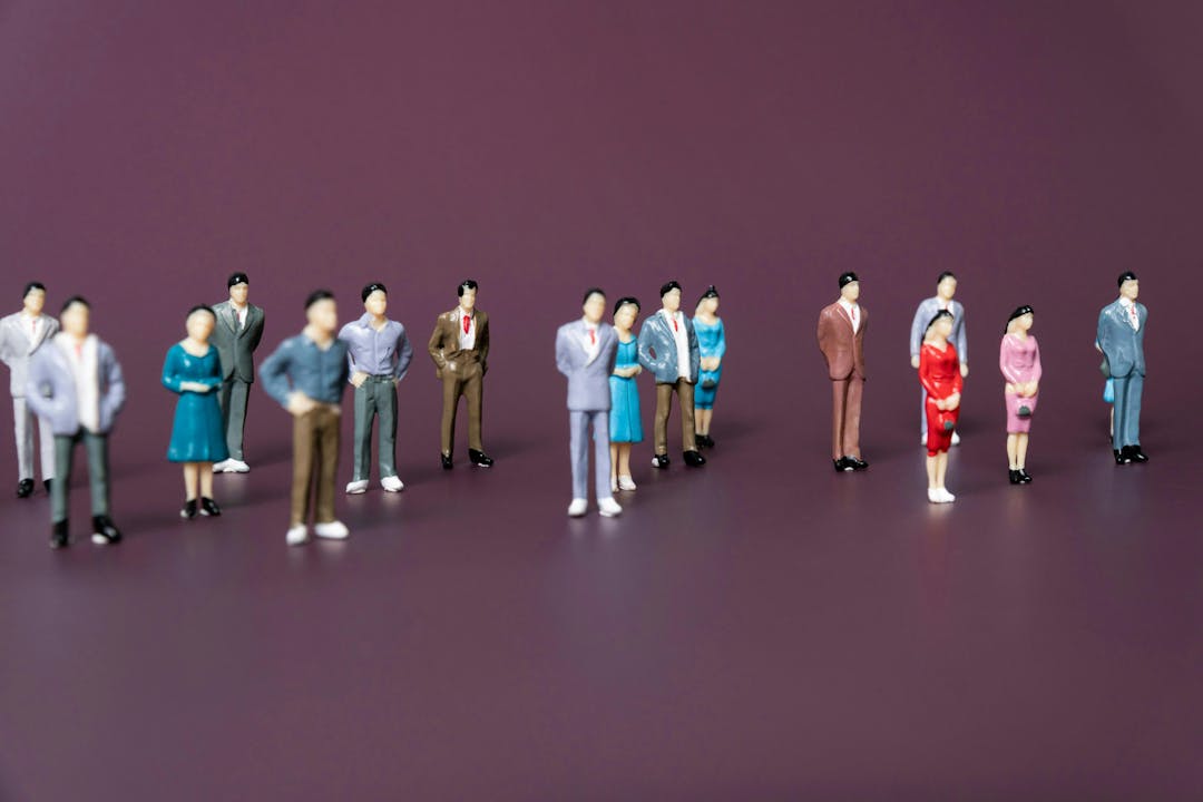 The image depicts a group of miniature figurines of people arranged on a plain, dark background. The figures are dressed in various formal and semi-formal clothing, including suits, dresses, and skirts. They appear to be standing still, with some looking straight ahead, others with hands on hips, and a few with arms crossed.