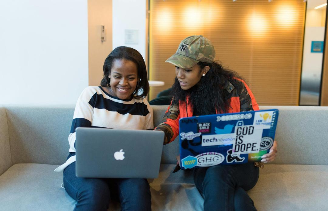 Two women sitting on sofa while using laptops.